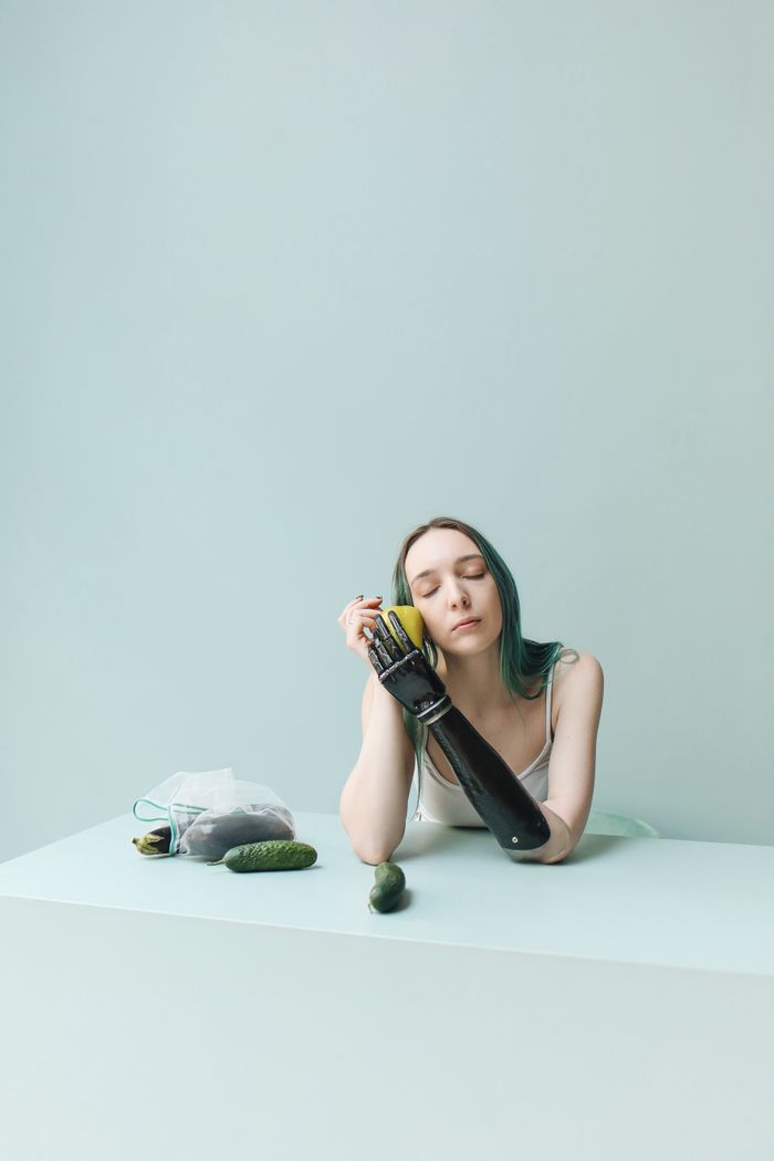 gallery-3 A young woman with a prosthetic arm holding a cucumber and apple at a pastel green table.