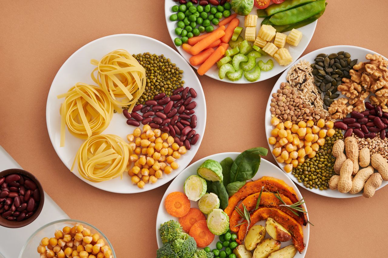 hero-img-01 A vibrant mix of legumes, vegetables, nuts, and pasta arranged on white plates showcasing a balanced, healthy diet.